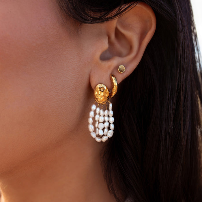 Close up of an ear waring unique textured gold earrings including a hammered gold oval with freshwater pearl drops, a hammered gold hoop, and an organic shaped hammered gold stud.