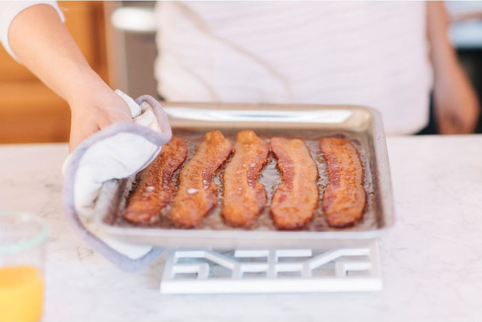 Front view of stainless steel jelly roll pan with four cooked bacon strips, showing the 1-inch lip and rim on a marble countertop