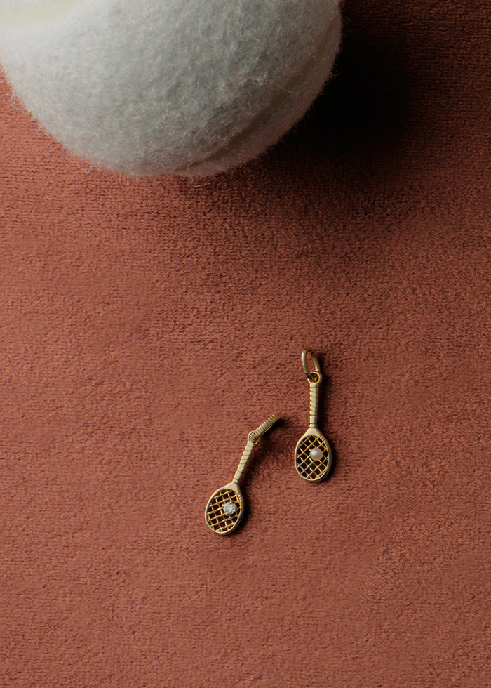 Two Kinn x Sunset Tennis Club 14k Gold Pearl Ball And Racquet Charms laying on a clay background next to a white tennis ball