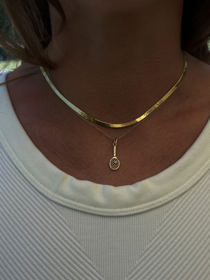A Kinn x Sunset Tennis Club 14k Gold Pearl Ball And Racquet Charm Necklace layered with a Carter Herringbone Chain Necklace