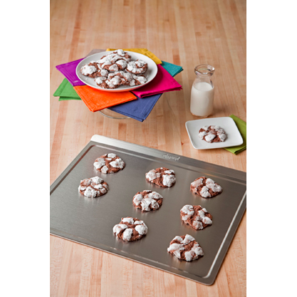 Angled view of large stainless steel cookie sheet showing rolled rim with cookies and a milk bottle on wood surface