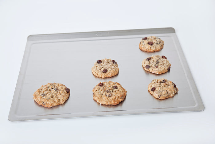Angled view of large stainless steel cookie sheet showing rolled rim and flat interior with six chocolate-chip cookies on white background