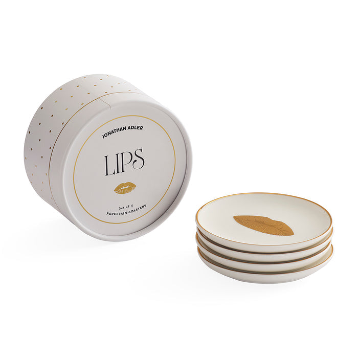 Jonathan Adler LIPS porcelain coasters, shown with the round branded storage container and a stack of four white coasters featuring gold rim detailing and a gold leaf motif.