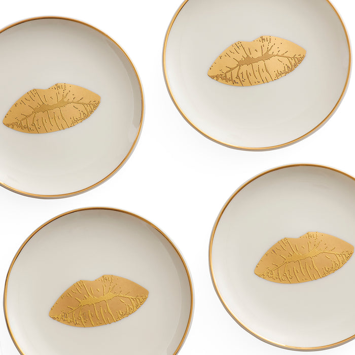 Four round white ceramic plates with gold rims, each decorated with a gold metallic lip motif in the center, arranged in a grid on a white background.