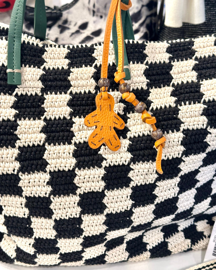 The Henry Beguelin Little Men Bag Charms feature a black and white checkered woven bag with green handles, yellow beads on one handle, an orange Italian leather gingerbread man-shaped charm, and a handmade logo.
