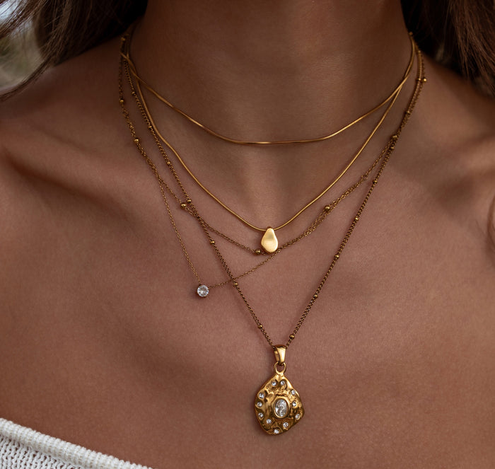 Close-up of a person wearing multiple gold necklaces with pendants on a neutral background