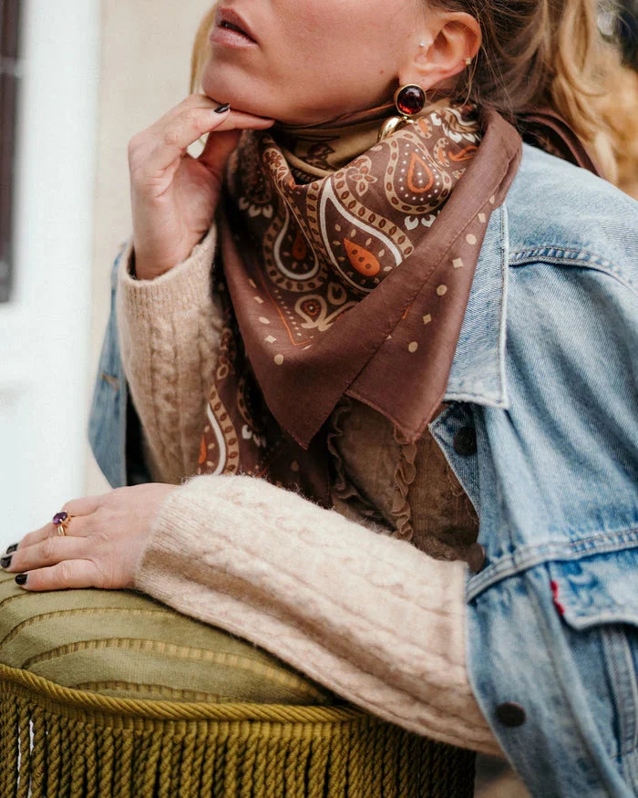 A woman wearing a beige cable-knit sweater, denim jacket, and the Maison Lecomte Flament Rodeo Scarf Cappuccino sits with her hand on a green cushion, showing off dark nail polish, statement earrings, and a ring.