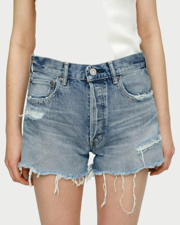 Close-up of a person wearing Moussy Packard Shorts by Moussy in a light blue wash, featuring frayed hems, multiple rips, and a button fly. Made from 100% cotton, these shorts have five pockets, including a coin pocket. The person is also wearing a white, sleeveless top.
