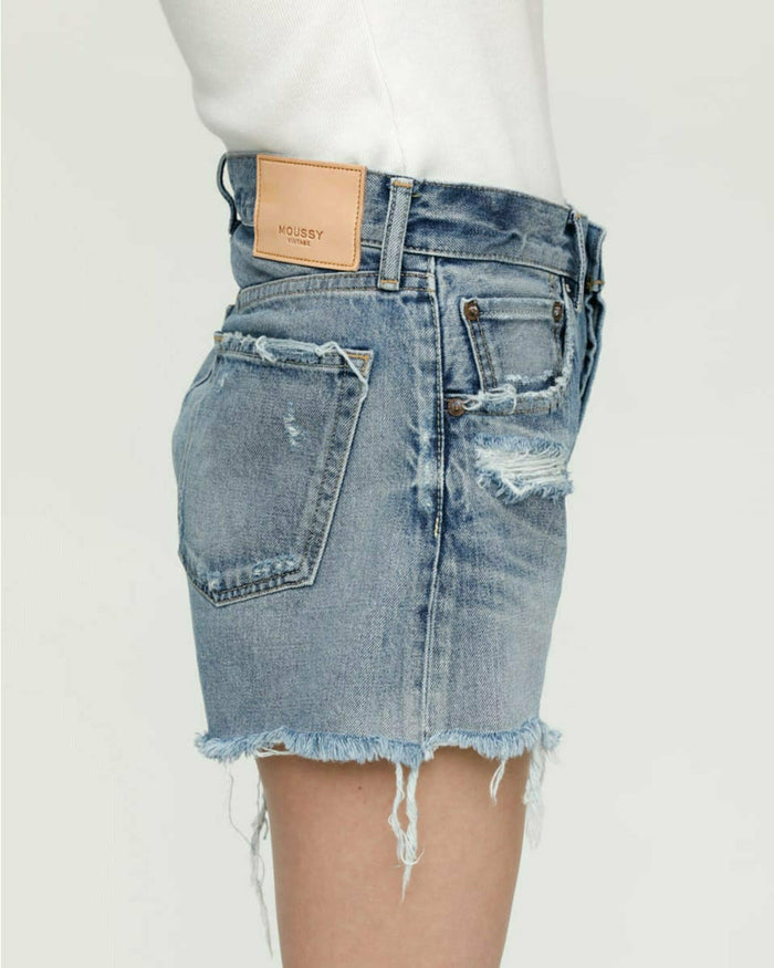 Side view of a person wearing light blue Moussy Packard Shorts by Moussy with frayed hems and a tan leather patch on the waistband with text. Featuring a button fly, these 100% cotton shorts are paired with a white top, but only the bottom portion is visible. The background is plain and light-colored.