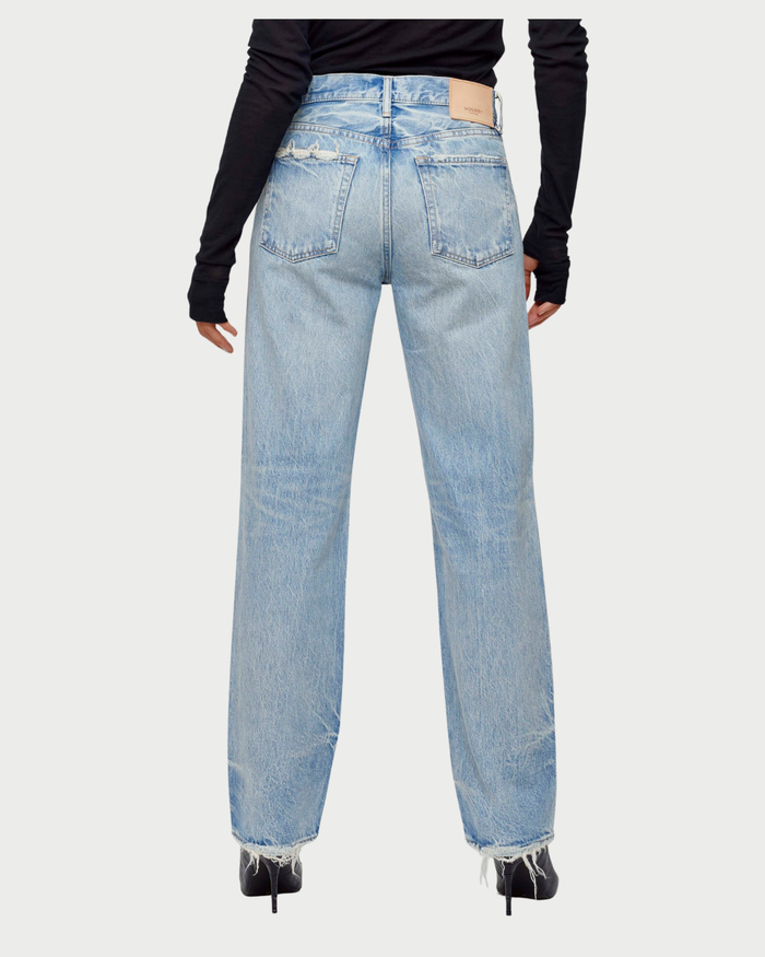 A person is seen from the back wearing a black long-sleeve top and Moussy Wildwood Straight Leg Jeans in light blue with frayed hems and distressed details, paired with black pointed-toe heels.