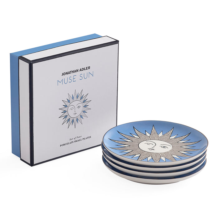 A set of four porcelain side plates with a blue and white sun design, shown next to their Jonathan Adler Muse Sun branded gift box on a white background.