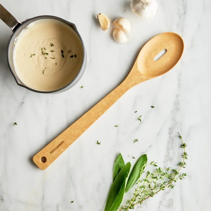 A light natural wood kitchen spoon with a handle cutout lies on a white marble surface next to a small pot of creamy sauce with herbs, garlic cloves, and green herbs.