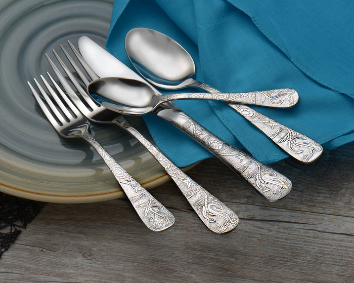 Close-up of Old Harbor flatware showing engraved decorative handles on spoons and forks on a ceramic plate with teal napkin