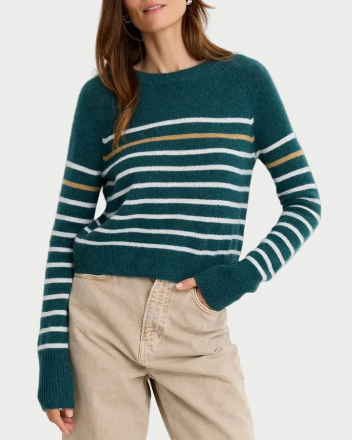 A woman wearing a dark teal knit sweater with horizontal white and mustard yellow stripes, paired with beige trousers, standing against a plain white background.