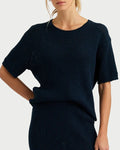 A woman models the One Grey Day Erica Knit Tee, a relaxed fit, lightweight knit with short sleeves and a round neckline, paired with a matching skirt. She poses with one hand on her hip against a plain background, face partially unseen.