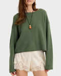 A woman modeling a sage green knit crewneck sweater paired with cream lace-trimmed shorts and a long pendant necklace, set against a plain light gray studio background.