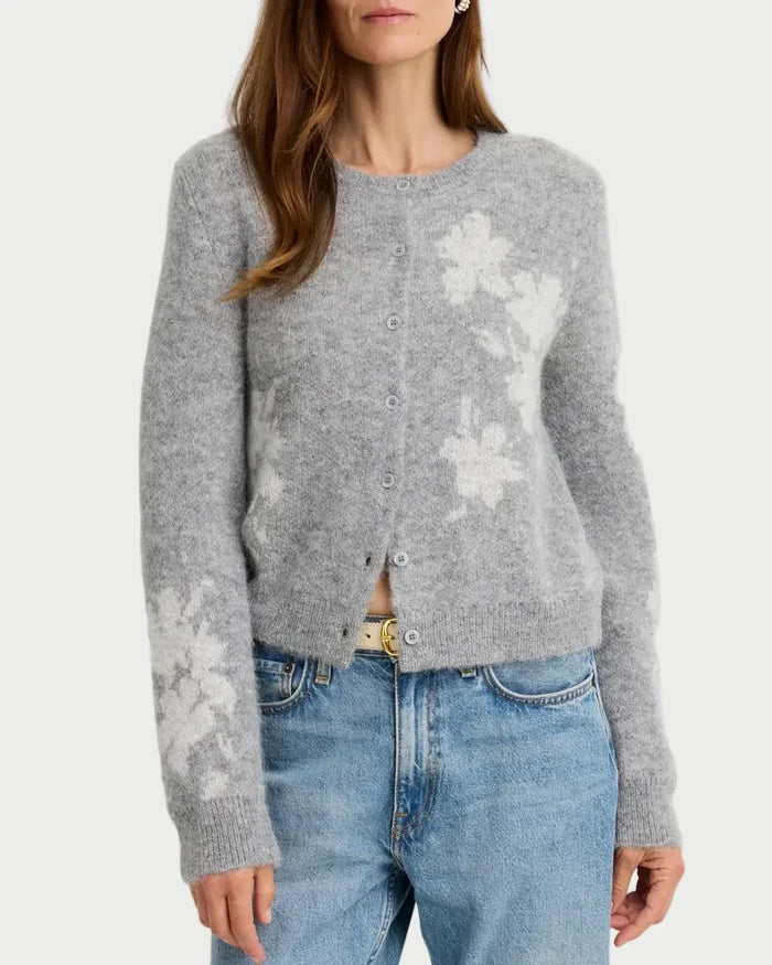 A woman wearing a light gray fuzzy knit cardigan with white floral patterns and light blue jeans, standing against a plain light gray background.