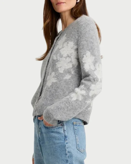 A woman stands in a studio wearing a light gray knit cardigan with a white floral pattern, light blue wide-leg jeans, and beige pointed-toe heels.