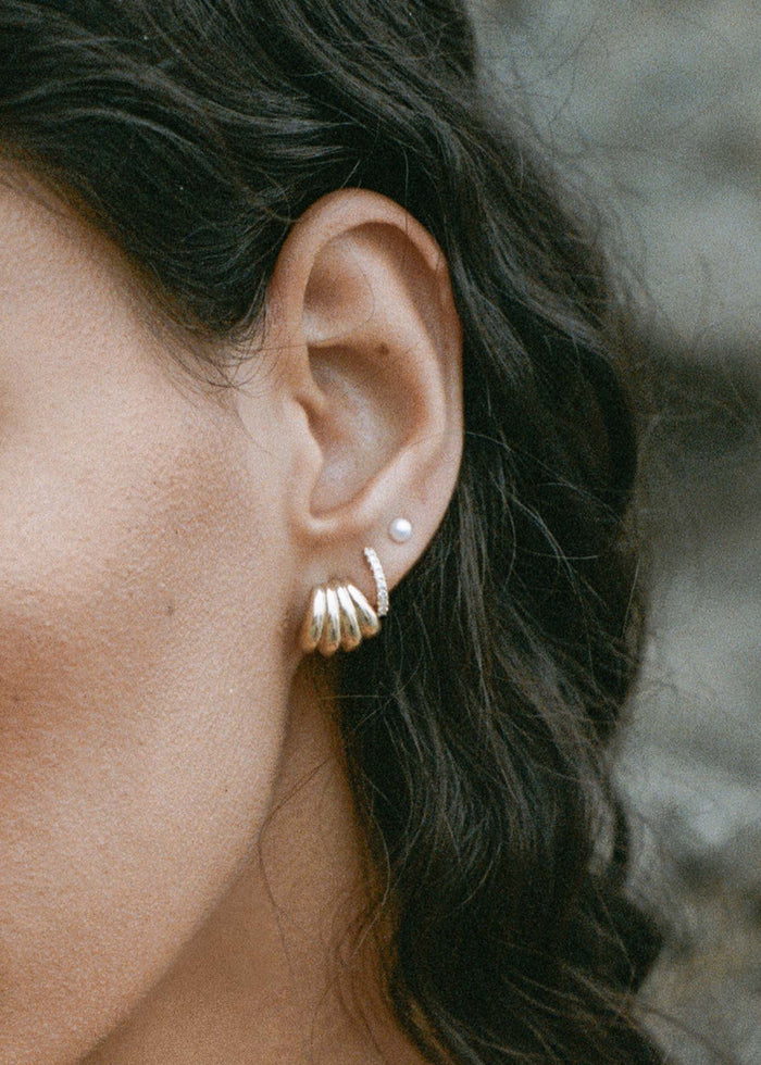 close up of a woman's ear wearing Kinn 14k Gold Gaia Scallop Earrings layered with Pave Diamond Huggie Earrings and a Pearl Stud