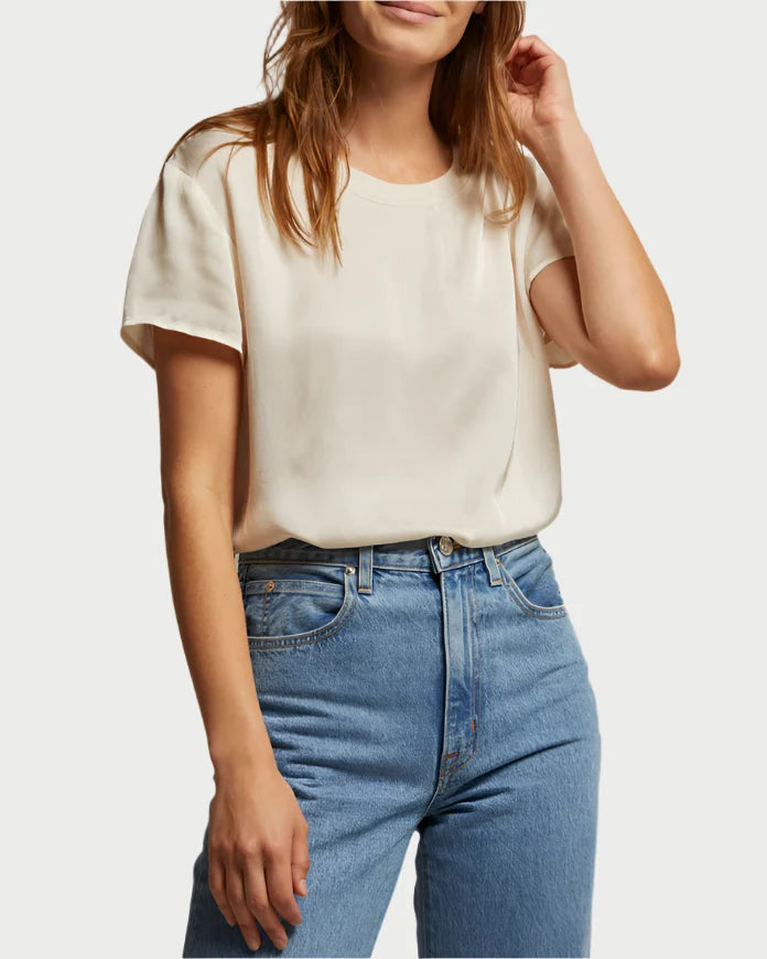 A woman wears the perfectwhitetee Delphine Satin Crewneck Tee in Sugar by perfect white tee, tucked into high-waisted blue jeans. She stands with one hand touching her hair and the other by her side; her face is partially visible.
