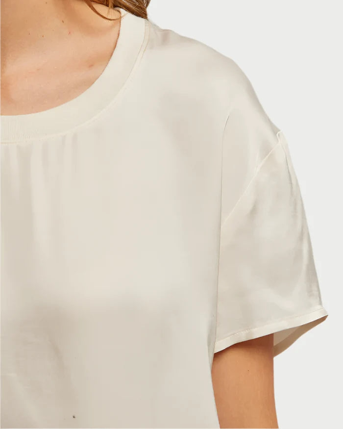 A close-up of a person wearing the perfectwhitetee Delphine Satin Crewneck Tee in Sugar by perfect white tee, highlighting the neckline, shoulder, and upper arm against a plain background.