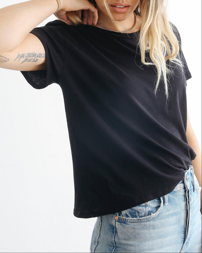 A woman with blonde hair and a forearm tattoo wears the perfectwhitetee Harley Boxy Crew in black by perfectwhitetee and light blue jeans, standing against a white background with one hand behind her neck; her face is partially out of frame.