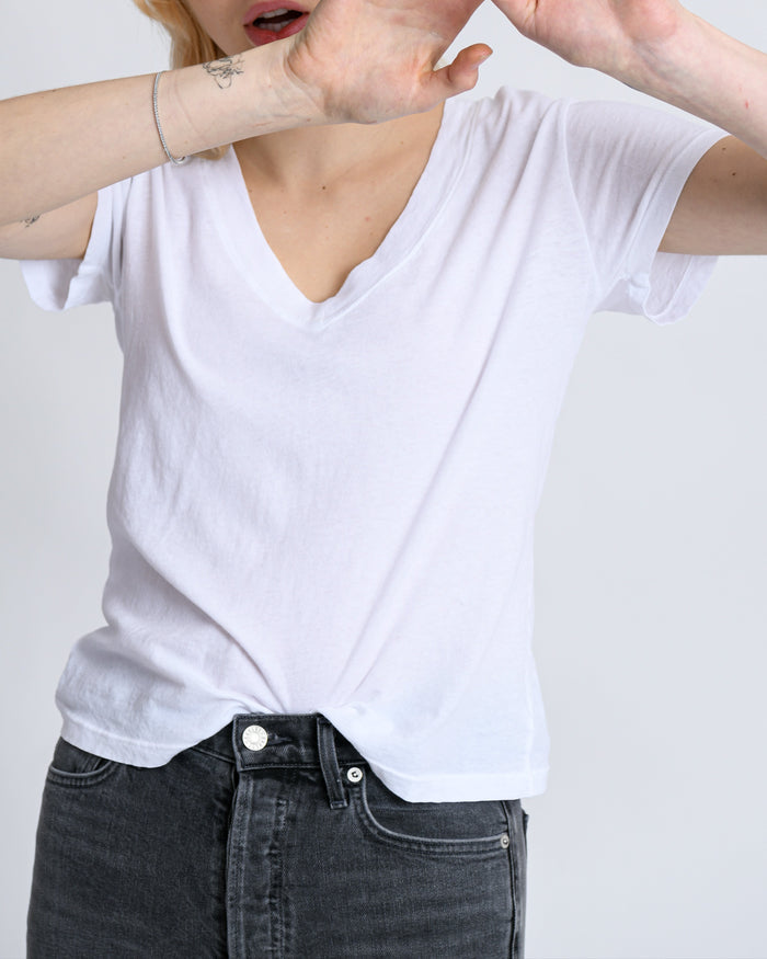 A person in a perfectwhitetee Hendrix Basic V-Neck T-Shirt in white and dark jeans stands against a light background, raising their arms to partly hide their face, with only the lower half visible.