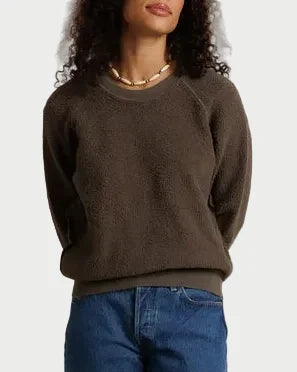 A woman stands against a draped off-white fabric backdrop wearing a dark brown crewneck sweater, blue jeans, and dark brown shoes.