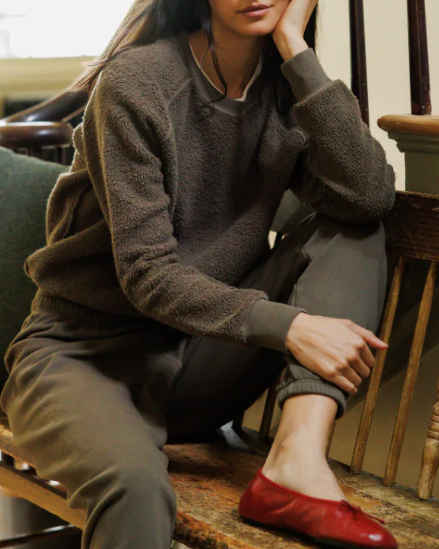 A woman sits on a wooden chair in a home interior, wearing a dark brown fuzzy crewneck sweatshirt, matching dark pants, and a single red flat shoe.