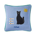 A square light blue throw pillow with a green piped edge, featuring an embroidered design of a black cat, three white fish, a blue wave, and a yellow star on a white background.