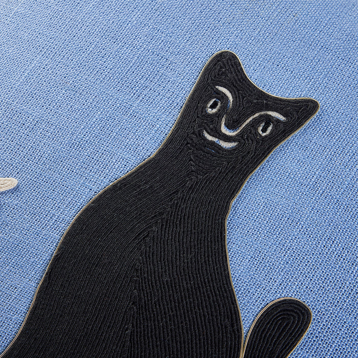 A close-up of a black embroidered cat patch with white facial details, placed on a textured light blue fabric background.