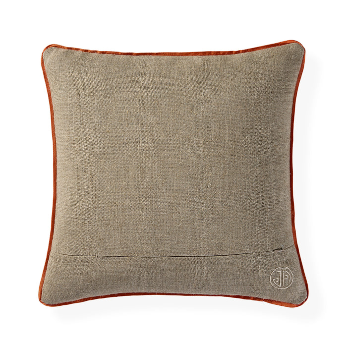 A square decorative throw pillow featuring a textured beige linen-like fabric body and a contrasting rust-orange velvet piped border, with a small circular embroidered monogram logo in the bottom right corner.