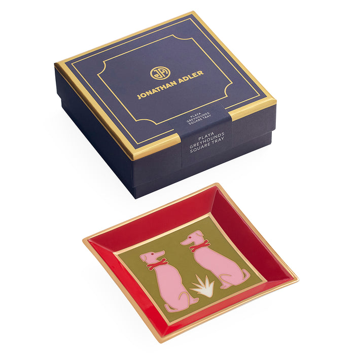 Jonathan Adler square glass dish with a red interior, gold trim, and pink dog illustrations, shown alongside its navy and gold presentation box on a white background.