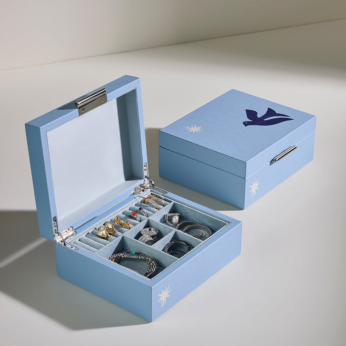 A light blue jewelry box shown open and closed on a neutral studio background. The open box contains multiple compartments holding rings and bracelets, and the closed box features a dark blue bird logo and small white starburst accents.