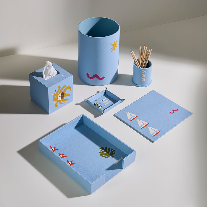 A coordinated light blue desk and bathroom accessory set arranged on a white surface, including a tissue box holder with a yellow sun, a cylindrical cup with a star, a small toothpick holder, a tissue box, a small tray, a rectangular tray, and a matching card or cover with sailboat motifs.