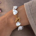 A gold chunky bracelet with white baroque pearls and a toggle closure, worn on a person's wrist.