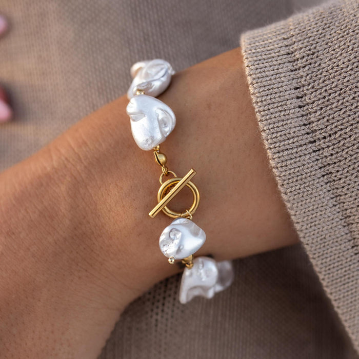 A gold chunky bracelet with white baroque pearls and a toggle closure, worn on a person's wrist.