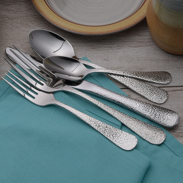 Set of textured silverware including two forks and two spoons resting on a teal cloth napkin, with part of a plate and a cup in the background on a wooden surface.