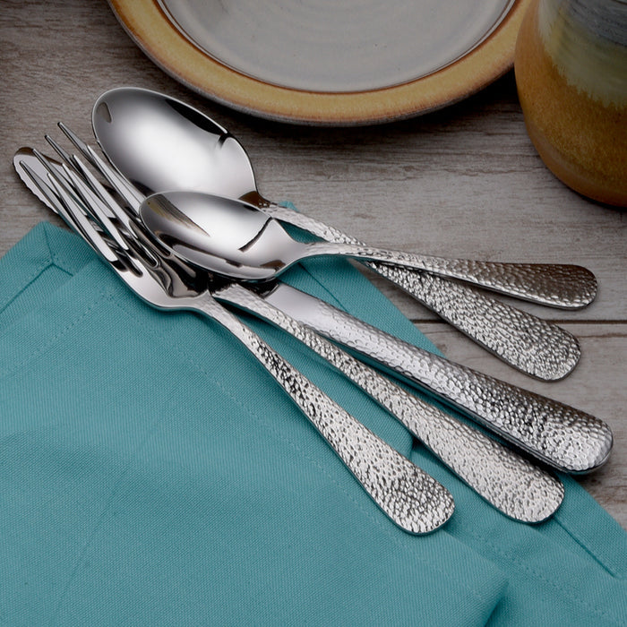 Set of textured stainless steel forks and spoons arranged on a teal cloth napkin with a plate and cup in the background