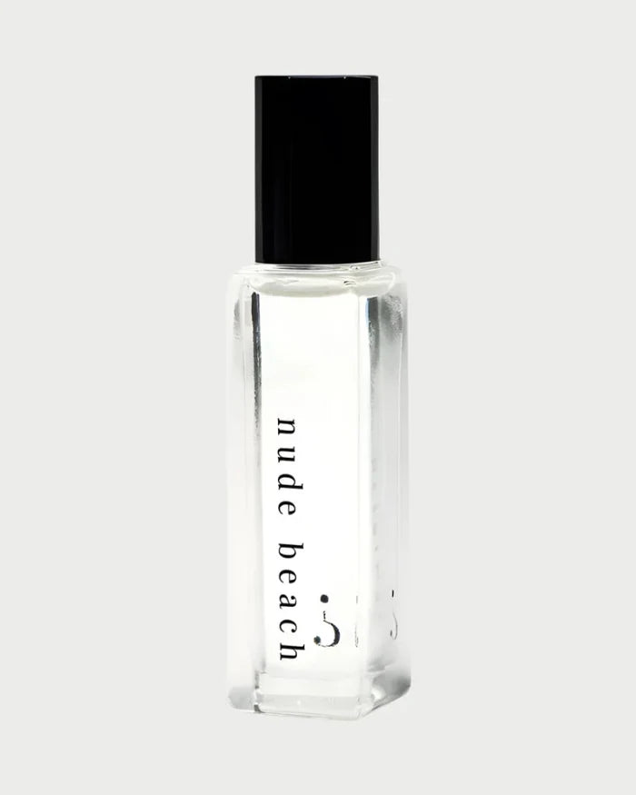A Riddle Oil - Nude Beach 8ml bottle, made of clear glass and sealed with a black cap, contains a transparent liquid. The label features the words "nude beach" in lowercase letters. The design is minimalist and sleek, showcasing the trending Nude Beach scent by Riddle Oil.