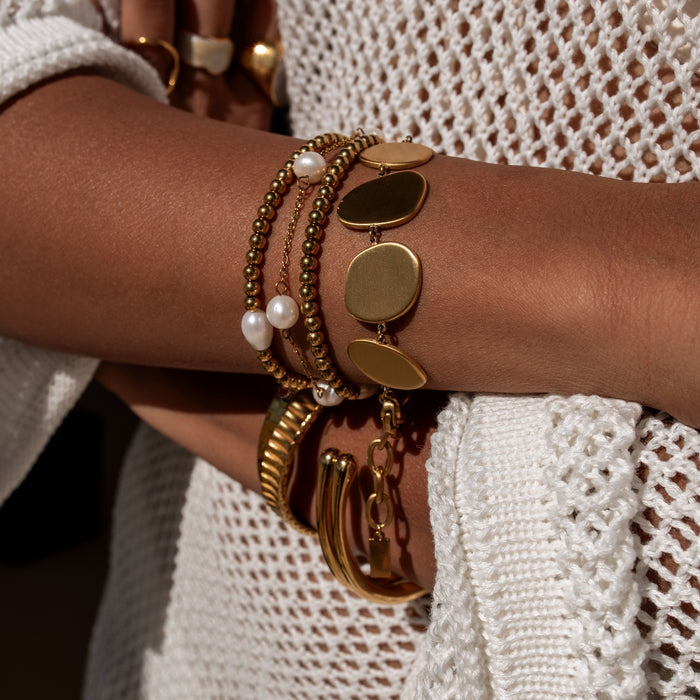 Close-up of a wrist wearing multiple gold bracelets with pearls against a white textured fabric background.