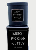 A black 9oz candle from Rude Dude with a label that reads "ABSO-F*CKING-LUTELY, scent of conviction" sits on a matching black box adorned with the same label. In the blurred background, a silver hand sculpture is visible, holding up its middle finger. Made in USA.