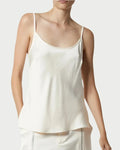 A person wears the Saint Art Elliot Ivory Charmeuse Camisole by Saint Art NY, a sleeveless white top with thin straps, paired with matching light-colored pants and shown from the waist up against a plain background.