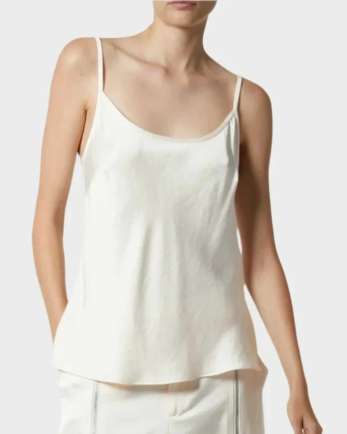 A person wears the Saint Art Elliot Ivory Charmeuse Camisole by Saint Art NY, a sleeveless white top with thin straps, paired with matching light-colored pants and shown from the waist up against a plain background.