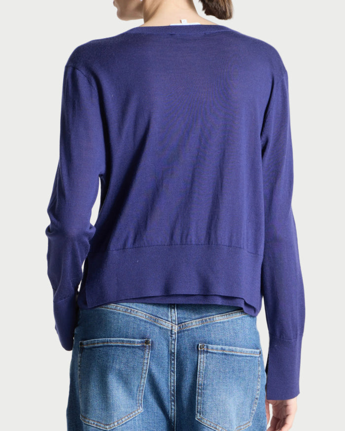 A person shown from the back wearing a cropped long-sleeve knit sweater in a medium periwinkle blue-purple color, paired with blue jeans, against a plain light gray studio background.