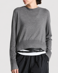 A person models the Saint Art NY Elodie Sweater—a loose gray merino wool piece—layered over a crisp white shirt, complemented by black pants and a black belt. Standing with one hand in their pocket against a plain background, they capture the effortless style of Saint Art NY.
