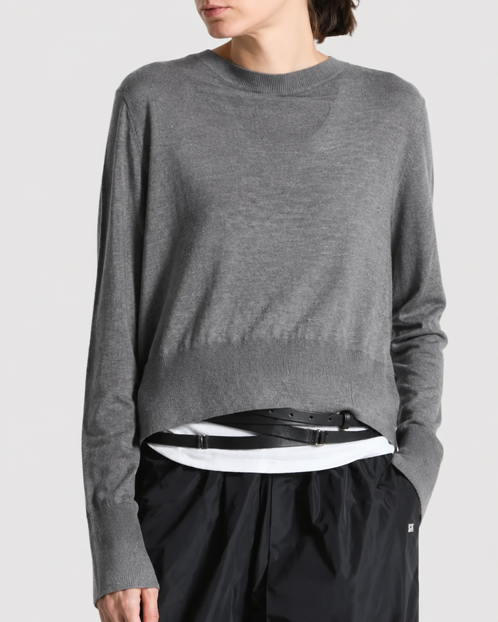 A person models the Saint Art NY Elodie Sweater—a loose gray merino wool piece—layered over a crisp white shirt, complemented by black pants and a black belt. Standing with one hand in their pocket against a plain background, they capture the effortless style of Saint Art NY.