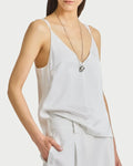 A person wears the Saint Art NY Kiera Tank, a white sleeveless V-neck top, paired with white pants and a long silver necklace, posing with one hand in their pocket against a plain white background.
