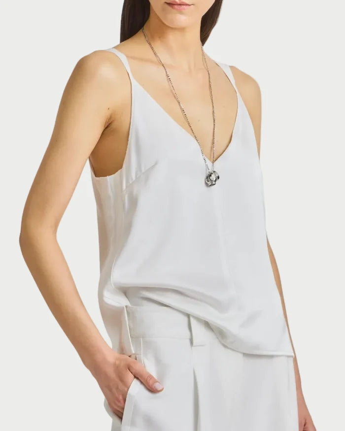 A person wears the Saint Art NY Kiera Tank, a white sleeveless V-neck top, paired with white pants and a long silver necklace, posing with one hand in their pocket against a plain white background.