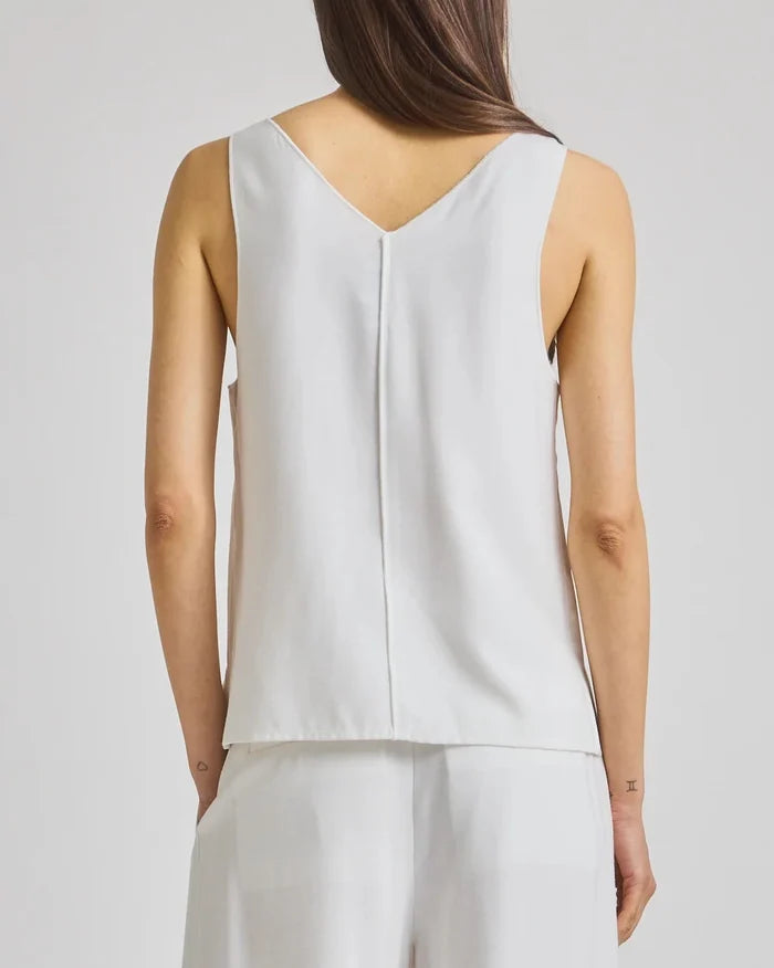 A woman faces away, wearing a sleeveless Saint Art NY Kiera Tank in white with a V-neck and matching pants. Her straight brown hair is down, arms relaxed at her sides, set against a plain neutral background.